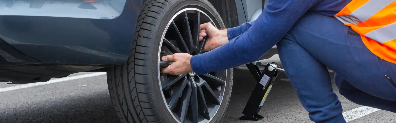 Can You Drive On A Flat Tire – Reasons And Solutions? A man in a orange safety vest changes a flat tire on a road. Close-up mans hands to the wheel of a